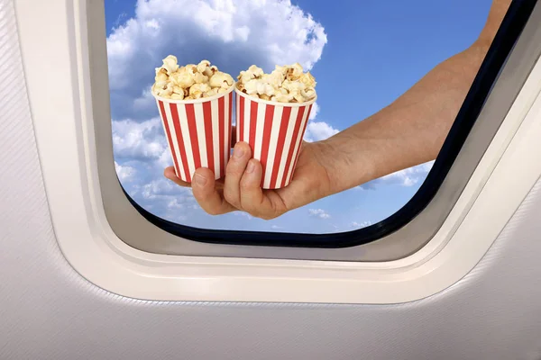Mano masculina sostiene dos tazas con palomitas de maíz sobre un fondo de un hermoso paisaje celestial de cielo azul con nubes esponjosas detrás del ojo de buey de un avión, concepto de viaje — Foto de Stock