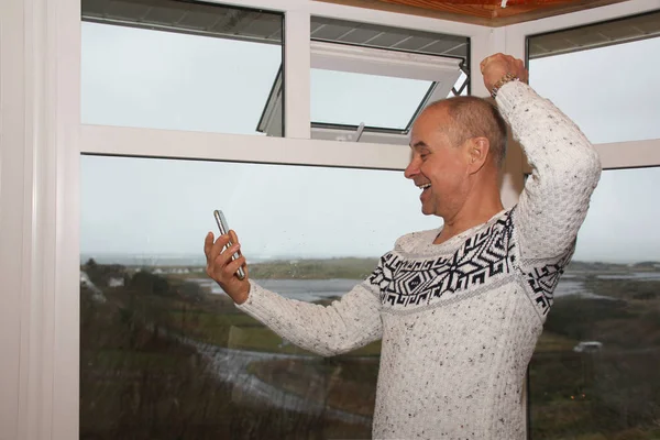Радостный мужчина в белом свитере стоит у широкого окна веранды и пользуется смартфоном, эмоционально улыбается — стоковое фото