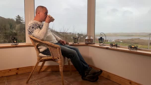 ベランダの居心地の良いウィッカーチェアに座って熱いお茶を飲んで窓から外を眺める男楽しさとリラクゼーションの概念 — ストック動画