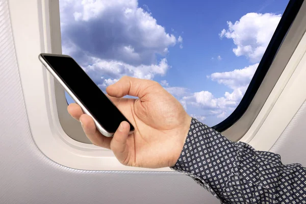 Mano masculina con un teléfono inteligente en el fondo de un hermoso cielo azul cielo paisaje con nubes esponjosas detrás del ojo de buey de un avión, concepto de viaje —  Fotos de Stock