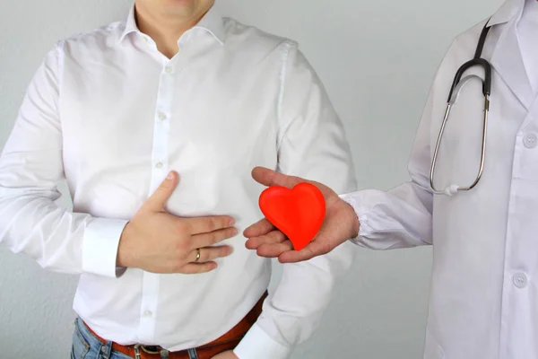 Hombre con una camisa blanca se aferra a su pecho en el área del corazón, a continuación es un médico con una bata blanca y un modelo de corazón rojo, concepto de problemas de salud, enfermedades del corazón, cirugía, trasplante de órganos — Foto de Stock