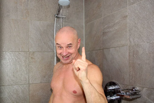 Пожилой человек моется в душе под ручьем воды и радостно улыбается, показывает предупреждающий жест рукой — стоковое фото