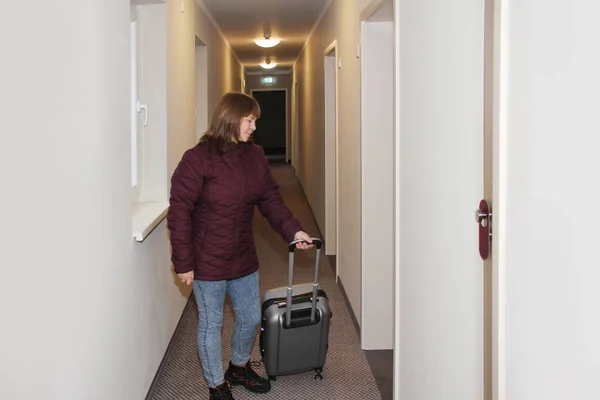 Горизонтальное фото женщины, туриста, идущего по длинному коридору отеля с чемоданом за дверью, туристическая концепция — стоковое фото