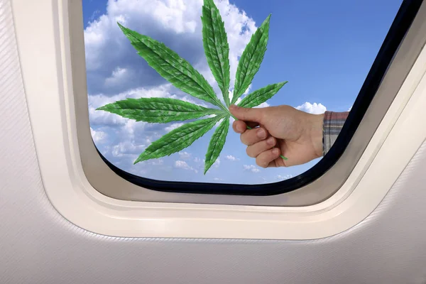 Mano masculina con una hoja verde de marihuana sobre el fondo de un hermoso paisaje celestial de cielo azul con nubes esponjosas detrás del ojo de buey de un avión, concepto de viaje — Foto de Stock