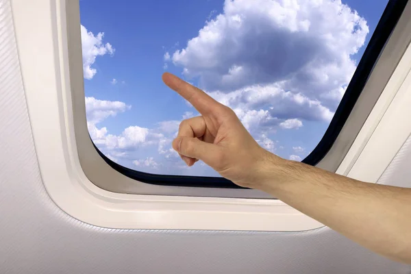 Mano masculina muestra en el fondo un hermoso paisaje celestial de cielo azul con nubes esponjosas detrás del ojo de buey de un avión, concepto de viaje —  Fotos de Stock