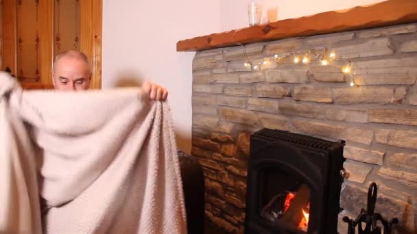 笑着的中年男人 裹着温暖的格子花 坐在靠近燃烧的壁炉的沙发上 冬眠的概念 — 图库视频影像