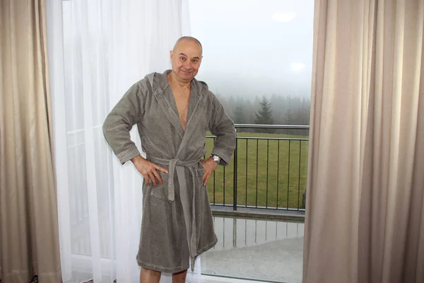 Iloinen keski-ikäinen mies harmaassa kylpytakissa seisoo hotellihuoneen ikkunassa, loma-käsite, matkailu — kuvapankkivalokuva