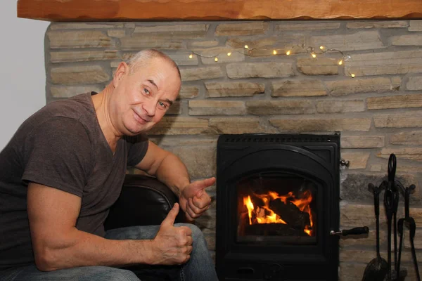 Happy middle aged man sitting near a hot burning fireplace, winter relaxation concept — Stock Photo, Image