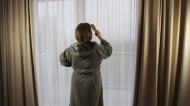 Gri bornozlu bir kadın otel odasının penceresinden saçını tarıyor, tatil konsepti, seyahat...