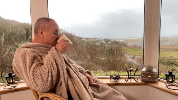 男人坐在阳台上温暖的毛毯里 坐在舒适的柳条椅上 喝着热茶 朝窗外看去 享受和放松的概念 — 图库视频影像
