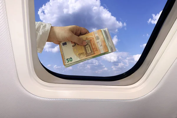 Avro banknotlarıyla dolu bir kadın eli uçağın lombozunun arkasında kabarık bulutlar ve seyahat konseptiyle mavi gökyüzünün güzel bir manzarasının arka planında.