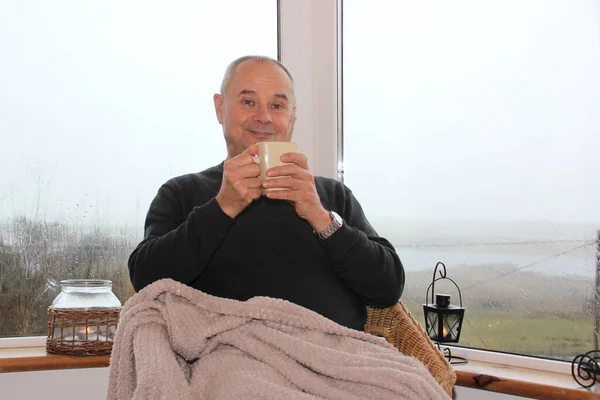 Homem senta-se em uma cadeira de vime aconchegante pela ampla janela na varanda, bebe chá, sorri, o conceito de descanso e relaxamento — Fotografia de Stock