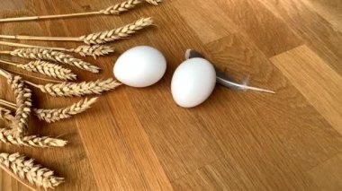 İki beyaz tavuk yumurtası ahşap bir masanın üzerinde duruyor, küçük bir tüy üzerine düşüyor, olgun buğday başaklarından oluşan bir arka plana.