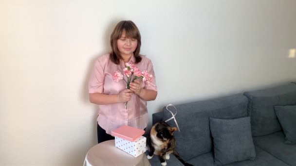 女人嗅着一束粉红色的康乃馨花 盒子里的礼物躺在一张圆桌上 一只黑猫坐在灰色的沙发上 舒适的家的概念 — 图库视频影像
