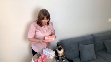 Kadın hediyeleri kutularda açar, bir buket pembe karanfil vazoda durur, siyah bir kedi gri bir kanepede oturur ve bakar, tatil konsepti, sevgililer günü, anne