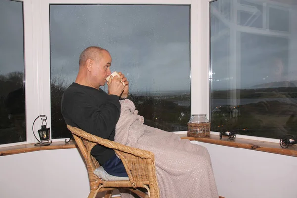 Homem senta-se em uma cadeira de vime aconchegante pela ampla janela na varanda, bebe chá, o conceito de descanso e relaxamento — Fotografia de Stock