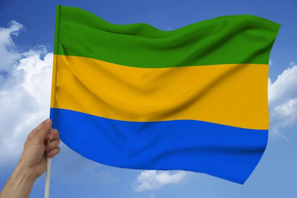 Hand houdt tegen de achtergrond van de hemel met wolken de kleur vlag van Gabon op de textuur van de stof, zijde met golven, close-up — Stockfoto