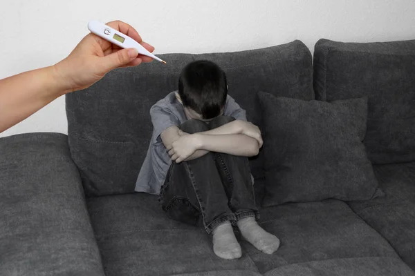 Niño triste, niño sentado en un sofá gris en la habitación y cubierto la cara con las manos, la mano de mamá extiende un termómetro, concepto de problemas de salud —  Fotos de Stock