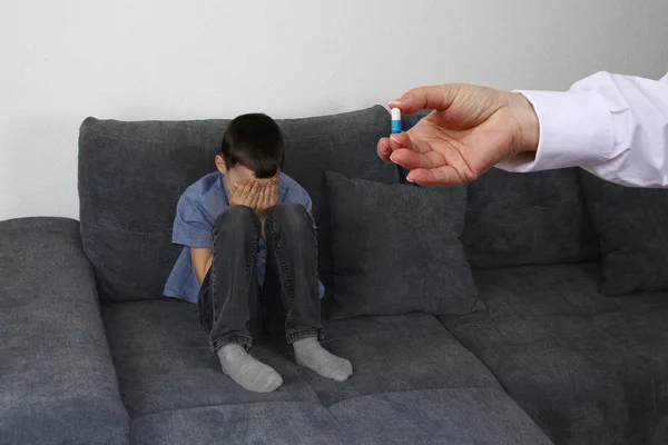 Niño, el niño se sienta en un sofá gris en la habitación y se cubre la cara con las manos, la mano de una enfermera o un médico extiende una píldora, el concepto de problemas de salud —  Fotos de Stock