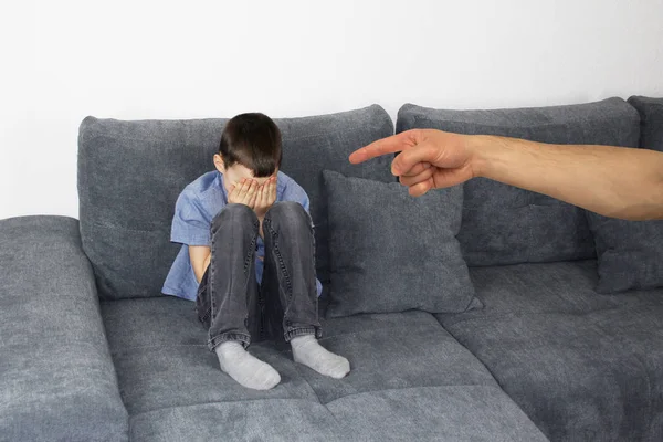 Trauriges Kind, ein Junge, der auf einem grauen Sofa im Zimmer sitzt und sein Gesicht mit den Händen bedeckt, die Hand des anklagenden Elternteils zeigt den Finger, das Konzept der schlechten Laune und der Erziehung — Stockfoto