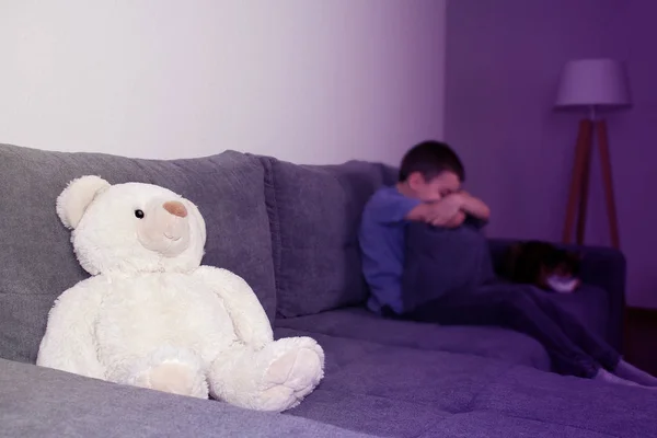 Niño, el chico en desenfoque escondió su cara en la almohada, un oso polar de peluche en foco se sienta en el sofá, el concepto de abuso psicológico, tintado púrpura —  Fotos de Stock