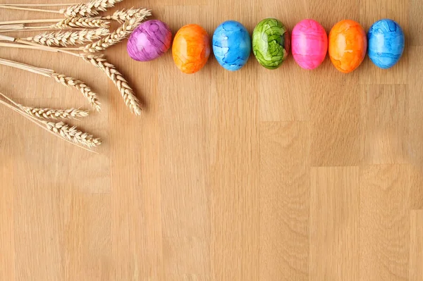 Вареные цветные яйца лежат на деревянном столе на фоне спелых колосьев пшеницы, Пасхальная концепция — стоковое фото