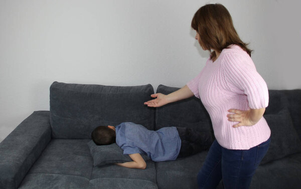 женщина, мать, ругающая сына, эмоционально жестикулирующая рукой, ребенок, лежащий с руками на диване, концепция семейного образования и конфликтов
