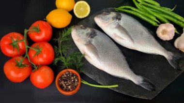 Limonlar, domatesler, acı biberler, iki taze çiğ Atlantik deniz balığı leşi Sparus aurata ve bir dereotu filizi siyah arka planda, yemek pişirme konseptinde uzanıyor.