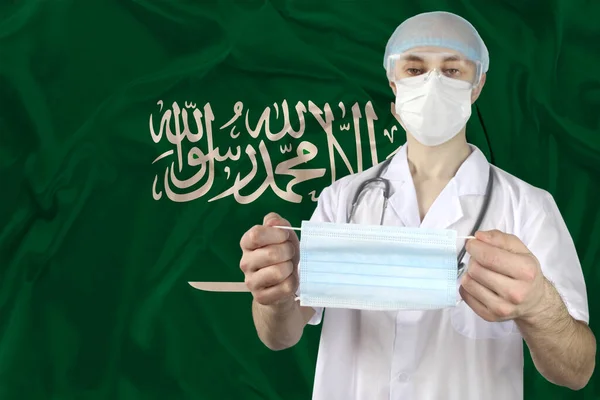 doctor in uniform with a stethoscope on the background of the silk national flag of Saudi Arabia is holding a medical mask, the concept of influenza, virus, COVID-19, coronavirus, treatment and vaccination