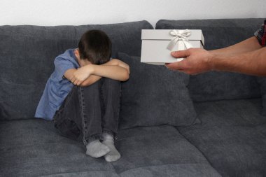 child, a boy sitting on a gray sofa in the room and covered his face with his hands, in the hands of a parent a gift in a box, the concept of children's vagaries and education clipart