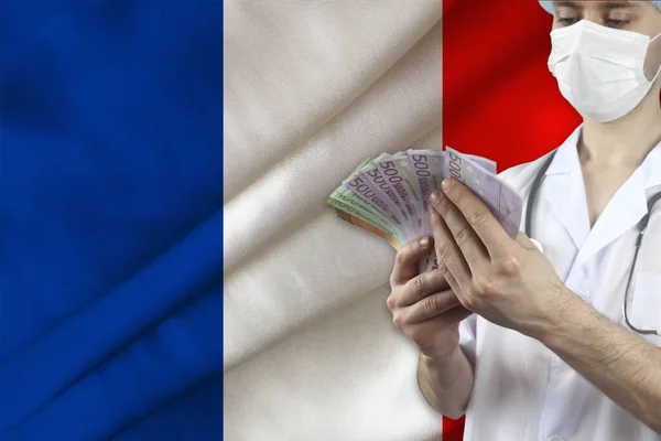 doctor with a stethoscope holds euro banknotes in the background of the silk national flag of France, the concept of medical insurance, budget, cost of treatment, organ donation
