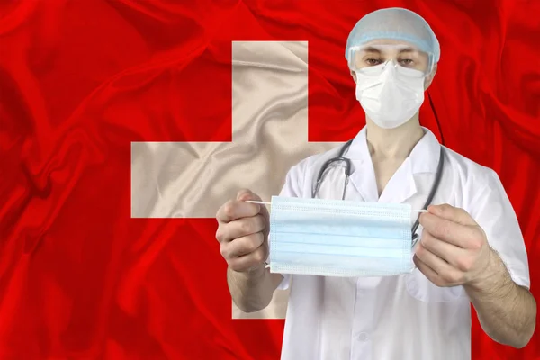 pharmacist, doctor holds a disposable protective mask against the background of the silk flag of Switzerland, the concept of personal protection against the SARS-CoV-2 virus, coronavirus, COVID-19, flu, infection, epidemic