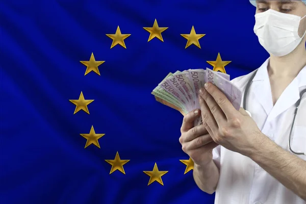 doctor with a stethoscope holds euro banknotes in the background of the EU silk national flag, concept of medical insurance, budget, cost of treatment, organ donation