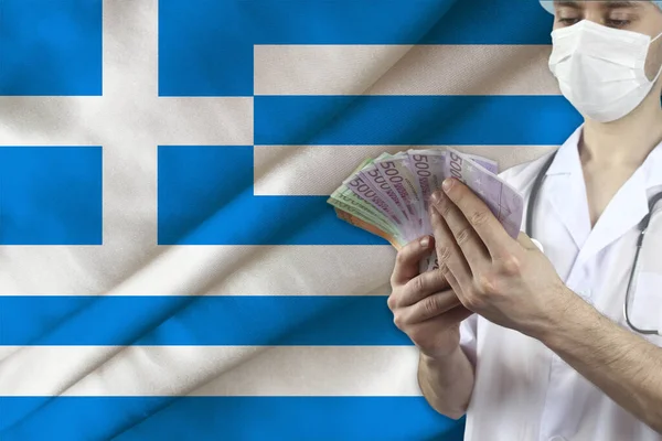 doctor with a stethoscope holds euro banknotes in the background of the silk national flag of Greece, the concept of medical insurance, budget, cost of treatment, organ donation