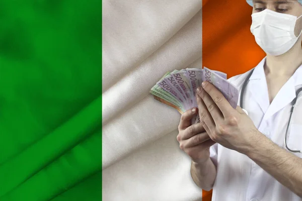 doctor with a stethoscope holds euro banknotes on the background of the silk national flag of Ireland, the concept of medical insurance, budget, cost of treatment, organ donation