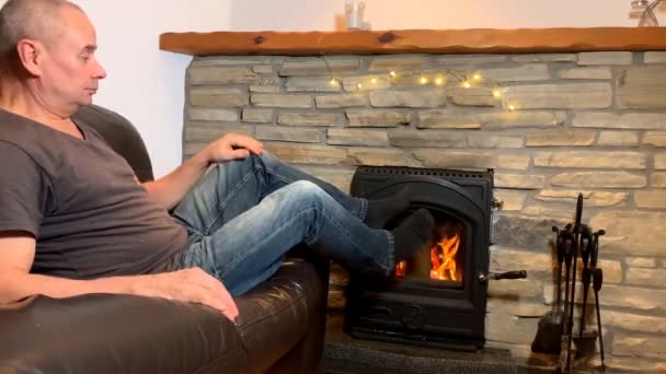 中年の男が燃える石で革のソファの上に座って温かさ 感情的に笑顔 居心地の良い家のコンセプトを楽しんでいます — ストック動画