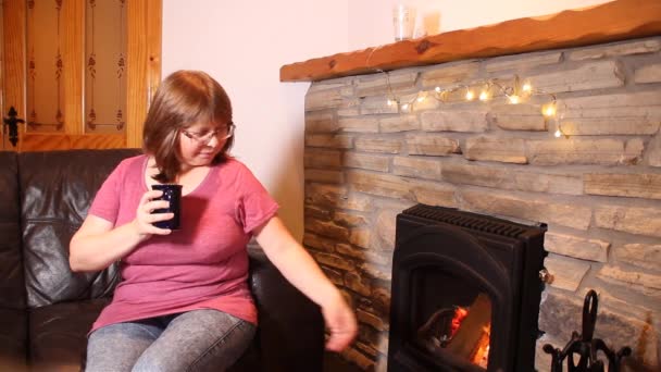 作为一个喝咖啡的女人 茶坐在一块燃烧的石头旁边的皮革沙发上 享受着温暖 亲切的微笑 享受着舒适的家的概念 — 图库视频影像