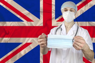 Büyük Britanya bayrağının arka planında bir doktorun elindeki tıbbi maske grip, Sars virüsü, Covid-19, koronavirüs, salgınlara karşı koruma kavramı.