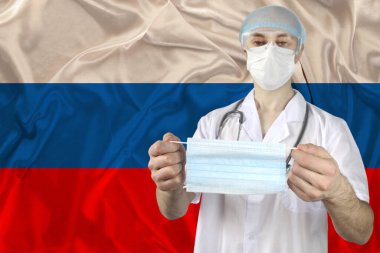 Rusya bayrağının arka planına karşı bir doktorun elindeki kişisel tıbbi maske grip, SARS virüsü, COVID-19, koronavirüs, salgınlara karşı koruma kavramı.