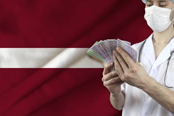 doctor with a stethoscope holds euro banknotes in the background of the silk national flag of Latvia, the concept of medical insurance, budget, cost of treatment, organ donation