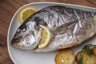 Sebzelerle pişirilmiş lezzetli Atlantik deniz balığı Sparus aurata limonlu beyaz bir tabakta yatıyor.