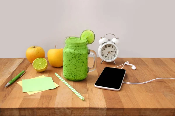 on a wooden table is a mason jar mug filled with useful smoothie of cucumbers, paprika, spinach, phone, alarm clock, apples, note paper, healthy eating concept, diet