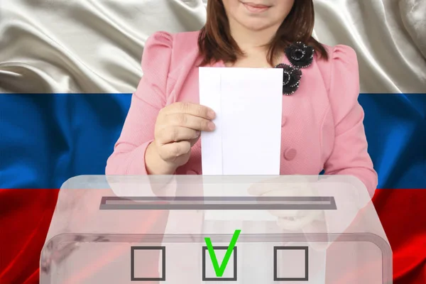 woman voter in a smart pink jacket lowers the ballot in a transparent ballot box against the background of the national flag of the country of Russia, concept of state elections, referendum