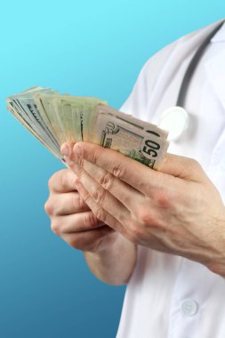 Koruyucu maskeli, steteskoplu bir erkek doktor elinde dolar banknotları, sağlık sigortası, bütçe, tedavi masrafları, organ bağışı kavramını tutuyor.