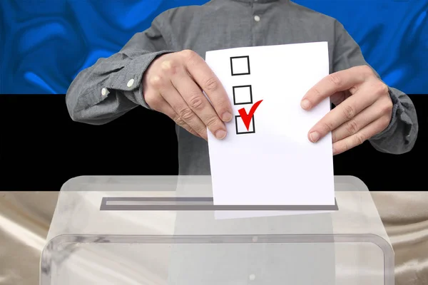 男性の有権者は エストニア国旗 国家選挙の概念 国民投票を背景に 透明な投票箱に投票を落とします — ストック写真