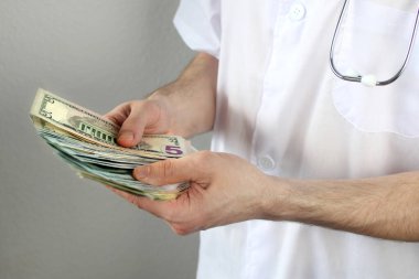 Koruyucu maskeli, steteskoplu bir erkek doktor elinde dolar banknotları, sağlık sigortası, bütçe, tedavi masrafları, organ bağışı kavramını tutuyor.