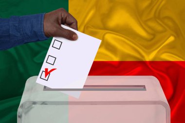 Erkek seçmen, Benin ulusal bayrağı, devlet seçimleri konsepti ve referandumun arka planında şeffaf bir sandık içinde oy kullanıyor.