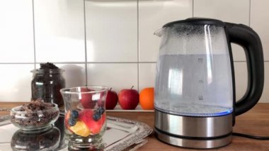 Kaynayan suyla modern cam çaydanlık, elma dilimleri, böğürtlen, dikenli çiçekler, arka planda meyveler, sağlıklı beslenme konsepti, vitamin kahvaltısı.