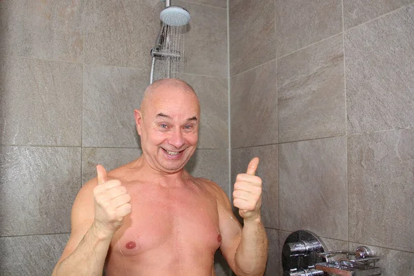 positive elderly man washes in the shower under a stream of water and smiles joyfully, shows gestures with his hands