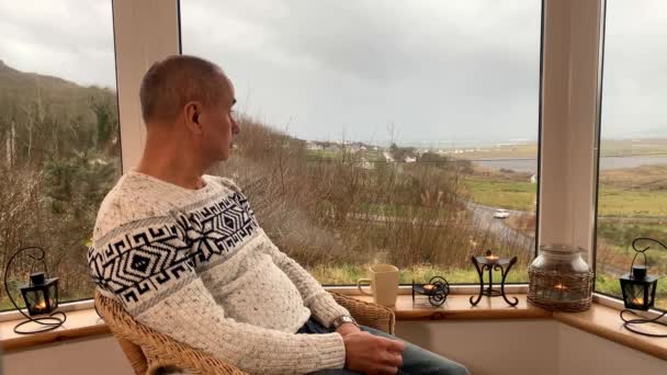 Ein Mann Sitzt Einem Gemütlichen Korbstuhl Neben Einem Großen Fenster — Stockvideo